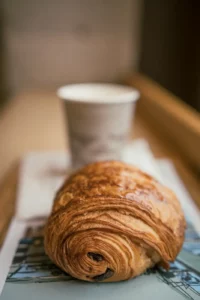 Photo d'un pain au chocolat devant un gobelet de café, sur un papier aux couleurs de la boulangerie de Yann Couvreur à Paris
